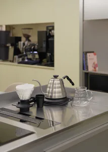 Stainless steel gooseneck kettle, glass pour-over dripper with paper filter, and glass server arranged on a reflective stainless steel countertop in a minimalist kitchen.