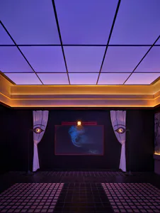 Dark surreal home theater room with deep purple coffered ceiling, warm cove lighting, two black eye-shaped floor lamps with white curtains, a large screen displaying blue abstract art, and a geometric purple velvet tiled floor.