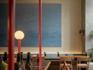 Modern restaurant interior featuring a large textured blue painting, built-in upholstered banquette seating, light oak wooden tables and chairs, and red structural poles in the foreground.