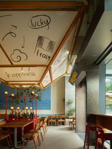 Modern restaurant interior with wooden ceiling structure displaying hand-drawn mural of lucky, Italy flag, happiness, and cloud motifs, red bar stools, wooden tables and chairs, and a blue wall with potted tree.