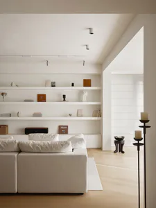 Modern minimalist living room featuring a large white sectional sofa, extensive floating white shelves displaying curated objects and books, light wood flooring, and simple cylindrical ceiling lights.