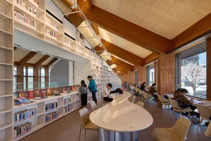 Modern library interior with tall white bookshelves, a large curved white marble island counter, wooden ceiling beams, and students studying at tables near large windows.