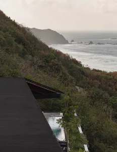 View from above of a modern villa with a dark timber roof and an infinity pool perched on a lush green cliffside overlooking the ocean and rocky coastline.