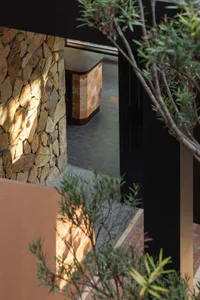 View through foliage of a sunlit natural stone wall and a modern entryway featuring terracotta patterned tiles and dark wood accents.