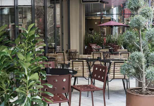 Outdoor dining patio of a sushi restaurant with red metal chairs, wooden slat tables, potted trees, and large glass windows in the background.