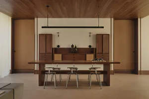 Modern minimalist dining room with a long walnut wood table, six round metal stools, wooden cabinetry, and a long black linear pendant light hanging from a wood-paneled ceiling.
