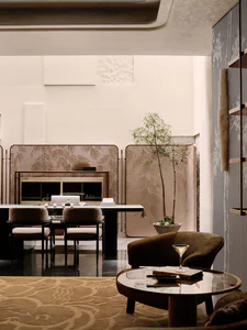 Elegant modern dining room featuring a long dark dining table with upholstered chairs, a marble console against a patterned folding screen, a tall potted bamboo tree, and a foreground seating area with a round coffee table and curved velvet armchair on a patterned rug.