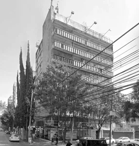 Black and white photograph of a tall concrete brutalist apartment building with multiple open balconies, surrounded by trees and overhead wires on a city street with pedestrians and cars.