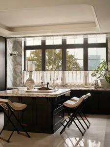 Modern luxury kitchen with a large black marble island, white marble walls, dark cabinetry, and three folding stools with cream cushions in front of a large window.