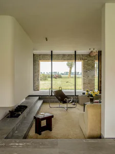 Modern minimalist living room featuring a built-in stone fireplace, brown leather sling chair, cream bouclé sofa, and large glass doors overlooking a green countryside landscape.