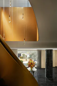 Modern double-height foyer featuring a curved golden brass wall, sculptural pendant lights, dark marble floor, and a large floral arrangement on a white table with a black marble column in the background.