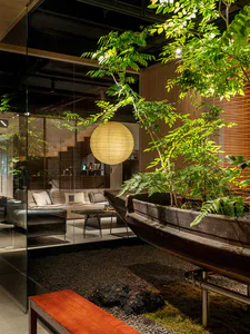 Modern living room viewed through glass with a large repurposed wooden boat used as a planter filled with ferns and small trees, paper lantern pendant light, dark leather armchair, and gravel floor in an indoor garden setting.