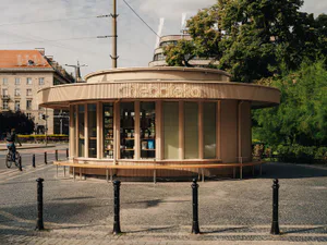 Modern circular wooden kiosk named Chleboteka with vertical ribbed cladding, large glass windows, and integrated seating bench on a sunny urban square in Prague.