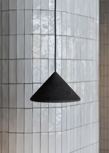 A black concrete cone-shaped pendant light hanging from a thin cord in front of a curved wall covered in small white rectangular tiles.