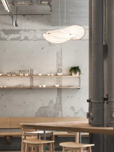 Interior view of a modern industrial-style cafe with exposed concrete walls, wooden tables and stools, floating wooden shelves displaying ceramics and plants, and a large sculptural white pendant lamp hanging from the ceiling.
