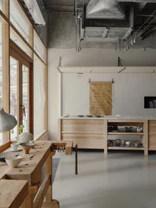 Modern minimalist cafe kitchen interior featuring light wood cabinetry, a wooden workbench in the foreground, exposed concrete ceiling with ductwork, and a large paper menu hanging on a white vertical slat wall.