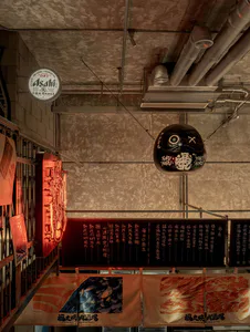 Low angle view of a Japanese yakiniku restaurant interior featuring exposed concrete ceiling, large black Daruma doll, Asahi beer sign, red neon, and hanging food banners.