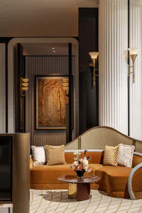 Luxurious living room featuring a curved burnt-orange velvet sofa with patterned and textured pillows, a round marble coffee table with floral arrangement, fluted white columns, and warm wall sconces, with a framed forest painting visible in the background hallway.