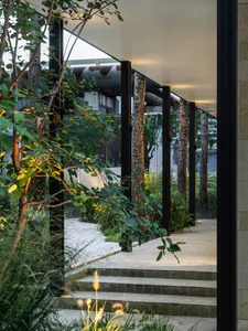 View from inside a modern building looking out through large glass windows and black frames into a landscaped courtyard featuring birch trees, grasses, and large weathered steel pipes under a white ceiling.