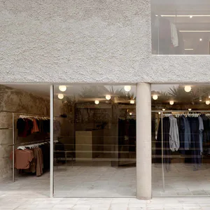 Exterior view of a minimalist boutique with textured plaster walls, large glass windows, clothing racks visible inside, and spherical pendant lights.