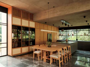 Modern dining room featuring a large teak wood table and six matching chairs, extensive built-in teak cabinetry with glass display, raw concrete ceiling, and large windows overlooking a lush tropical garden.
