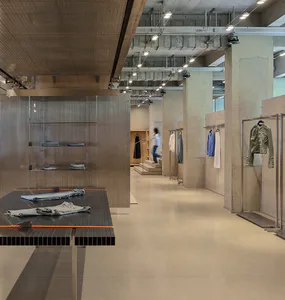 Modern industrial-style clothing boutique interior with raw concrete walls, polished concrete floor, metal mesh display shelves, and dark slatted display tables holding folded denim garments.
