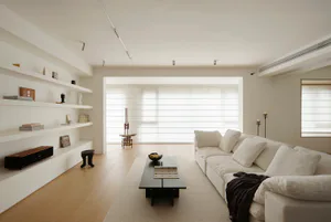 Bright minimalist living room featuring a large white sectional sofa, dark coffee table on a light rug, built-in white shelves with decorative objects, light oak flooring, and a large window with sheer horizontal blinds.
