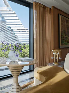 Modern luxury living room interior featuring a sculptural light-wood pedestal table with purple flowers, mustard yellow curved sofa, walnut wall paneling, and a dramatic city skyscraper view through floor-to-ceiling windows.