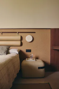 Modern minimalist bedroom corner showing a beige leather headboard with cylindrical bolsters, patterned and checkered pillows, a striped ottoman used as a nightstand holding a glass of water and a book, a circular wall sconce, and warm textured walls with wooden accents.