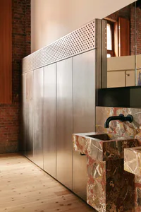 Modern industrial-style bathroom featuring a solid marble sink with black wall-mounted faucet, paired with tall brushed stainless steel cabinetry that has a perforated metal band along the top, set against exposed red brick walls and light wood flooring.