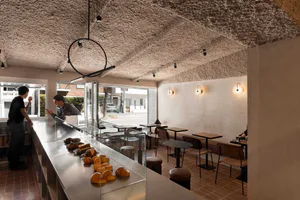 Interior view of a modern bakery cafe with a long stainless steel and glass pastry display counter filled with croissants and pastries, two men interacting at the counter, wooden tables and chairs on the right, and a distinctive textured plaster ceiling with circular pendant lighting.