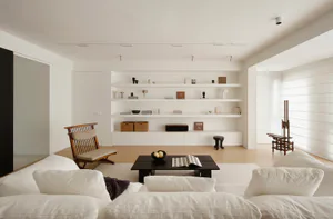 Modern minimalist living room with white L-shaped sofa, dark wood coffee table, wooden lounge chair, and extensive built-in white shelving filled with books and decor.