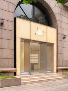 Exterior view of a modern boutique entrance featuring warm travertine stone cladding, a large arched window, glass pivot doors, and minimalist signage for 'yeye'.