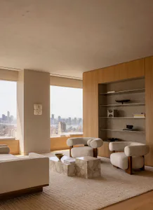 Modern minimalist living room interior with two boucle armchairs, marble coffee tables, built-in oak shelving, and large windows overlooking a city skyline.