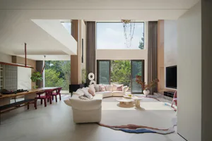 Modern double-height living room with white curved sectional sofa, marble floor, large windows overlooking green trees, and a colorful abstract rug.