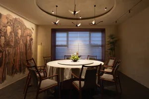 Elegant private dining room with a large round table covered in white linen, surrounded by eight walnut wood armchairs with grey upholstery. A large Tang dynasty mural depicting court ladies in traditional robes covers the left wall, while a floor-to-ceiling window with sheer blinds reveals a twilight city view. Sculptural red, black, and white bird-shaped pendant lights hang from a circular recessed ceiling above a central floral arrangement.