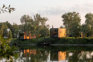 Two modern floating cabins with large glass windows and corten steel cladding situated beside a calm lake surrounded by lush green trees at sunset.