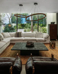 Modern living room featuring a large white sectional sofa, two dark brown leather armchairs with wooden frames, a black coffee table with geometric pattern on a jute rug, and two large interlocking circular wooden pendant lights hanging from the ceiling. Large windows reveal a green garden outside.
