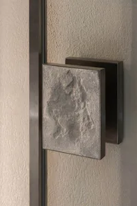 特寫顯示方形粗獷紋理混凝土門把手，表面有機自然紋理，安裝在深色金屬固定件上，背景為質感牆面。