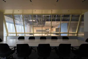 Modern conference room with black office chairs around a long white table facing a large golden-framed glass partition wall that reveals the ceiling infrastructure and adjacent lounge area.