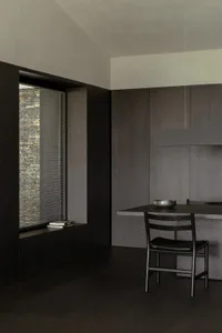 Minimalist dark wood dining area featuring a built-in window seat with horizontal blinds overlooking a stone wall, paired with a sleek dark wood dining table, matching chair, and metallic bowl under a linear pendant light.