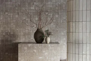 A modern interior corner with grey and beige patterned ceramic tiles on the wall and a pedestal, featuring a large dark ceramic vase with twisted dried branches and a small white vase with pine and red berries, next to a curved tiled column.
