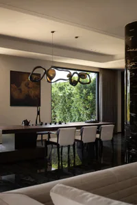 Modern dining room featuring a long dark wood table, four white upholstered chairs, a large abstract sculptural chandelier, dark marble floor, and large windows overlooking lush green trees.