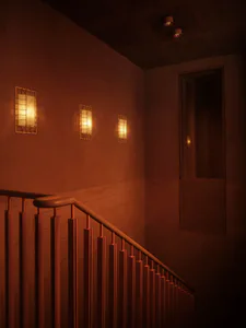 Interior view of a concrete staircase with wooden handrail descending into a warmly lit basement corridor featuring three illuminated wall sconces and recessed ceiling lights.