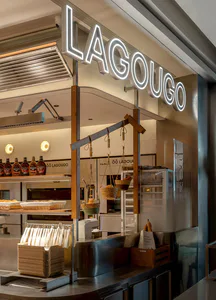 Lagougo 現代麵包店吧台，大型白色霓虹招牌、木質結構橫梁、懸掛藤編麵包籃、不鏽鋼檯面與溫暖照明。