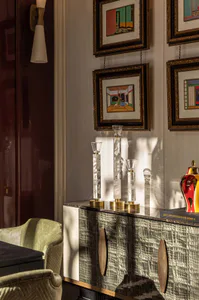 A luxurious dining room corner featuring a crocodile-embossed green sideboard topped with mother-of-pearl candle holders, colorful framed art on the wall, a modern wall sconce, and green velvet dining chairs.