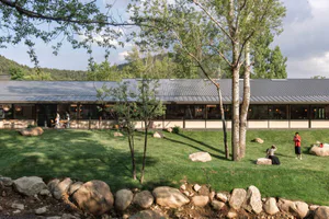Modern single-story resort building with dark corten steel roof, large glass windows, and a spacious green lawn dotted with natural boulders and birch trees under a clear blue sky.