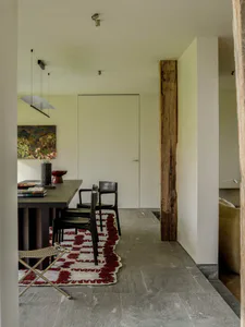 Modern dining room featuring a large reclaimed wood beam, dark wooden dining table with black chairs, red and white patterned rug on grey stone floor, and contemporary pendant light.