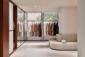 Modern minimalist clothing boutique interior with terrazzo floor, wooden wardrobe, black clothing racks filled with neutral-toned coats, and a curved beige fabric sofa in front of large windows overlooking trees.