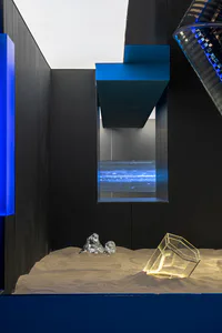 Modern art installation featuring a sand-covered base, metallic rock-like sculptures, a glowing transparent acrylic cube, and a digital blue light panel set within black geometric walls with blue accents.